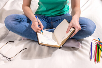 A girl in a green T - shirt and blue jeans is sitting on the bed and writing in a notebook , colored pencils and glasses are scattered around
