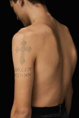 Naklejka premium Close-up shot of a young man with a water-based temporary cross tattoo on his shoulder. The portrait of a shirtless tattooed young man standing on a dark background. Back view.