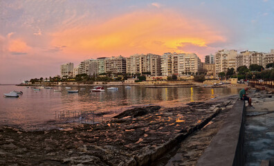 Sliema, Malta -  A man sitting on a wall at the rocky Exiles Bay Beach with a golden sky above...