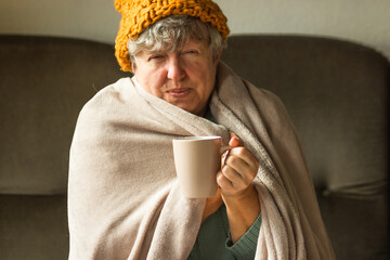 Elderly woman in ginger cap wrapped in blanket and drinking cup of tea. Old grandma feeling cold at home. Energy crisis.