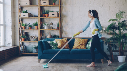 Fototapeta na wymiar Cheerful young woman is dancing with mop during routine clean-up in nice loft style apartment, she is listening to music with wireless headphones and singing enjoying her occupation.