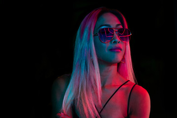 Asian young woman's portrait on dark studio background in neon wearing sunglasses in neon. Concept of human emotions, facial expression, youth, sales, ad.
