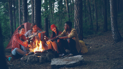 Male tourist is playing the guitar sitting near campfire with friends singing and having fun, young people are holding sticks with marshmallow above flame. Food, music and fun concept. - 541694156