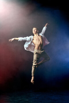 Male ballet dancer making dance jump element in smoke fume on isolated black background with red and blue light. Young man in jeans and bare naked torso