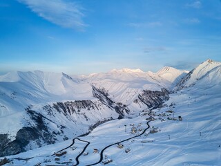 Obraz premium Wide aerial panorama of snowy mountain ridge at ski resort village on sunrise. Stunning mountains range covered with snow powder on ski resort at sunset. Caucasus mountain peaks skyline on sunset.