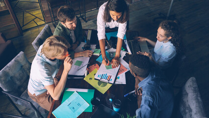 Multiracial workgroup is analyzing diagram and writing on colored stickers sitting at desk in office room. High angle view of people talking, pointing at charts and taking notes.