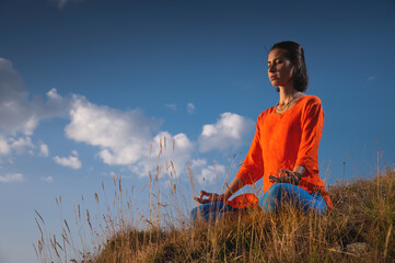 young caucasian woman meditates in the lotus position in the grass in the mountains near the cliff...