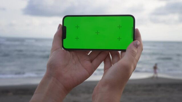 Point of View of woman holds horizontal mobile phone two hands and watching a video on background of ocean beach and walking people. Use green screen for copy space. Chroma key mock-up on smartphone. 