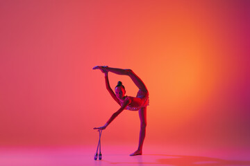 Emotions in motion. Portrait of junior female gymnast in black sport swimsuit doing gymnastics excercises isolated over pink background. Sport, skills, achievements