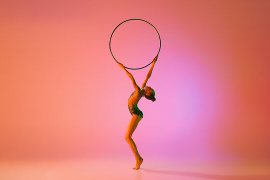 Emotions in motion. Portrait of junior female gymnast in black sport swimsuit doing gymnastics excercises isolated over pink background. Sport, skills, achievements