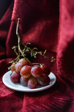 Bunch of grapes on a white plate against background of red luxury fabric. Vertical photo with grapes on the background of blurred red textile