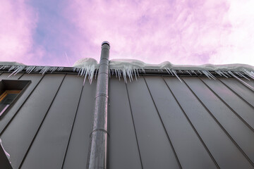 Frosted chimney pipe and icicles hanging from eaves of roof on cold day in dramatic sky background,...
