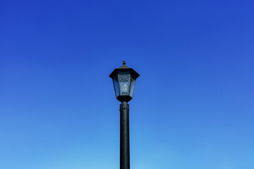Fototapeta na wymiar Dirty black lamppost with an electric bulb against a bright blue daytime sky. Horizontal background with sky and vintage city electric lantern in close-up