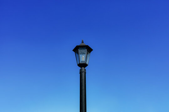 Dirty black lamppost with an electric bulb against a bright blue daytime sky. Horizontal background with sky and vintage city electric lantern in close-up
