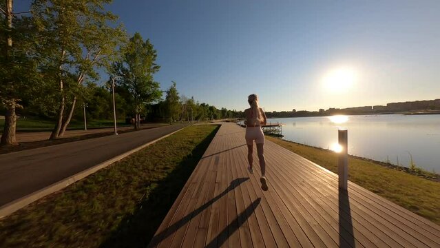 Aerial FPV drone view of sporty young woman in activewear running along wooden pier around lake on bright sunny summer morning. Back tracking view of running female active training in park.