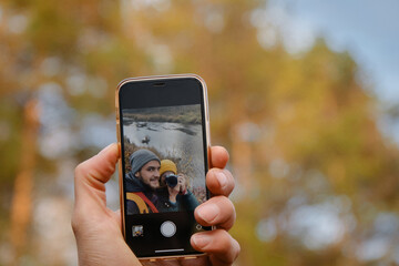 Walk near river, shoot video for travel blog. Young Caucasian couple in love takes selfie on smartphone while hiking in nature. Female wildlife photographer with camera.