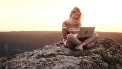 Cinematic shot of charming young woman have distance work or study online or writing a book sitting on the rock in the mountains with beautiful pink sunset. 