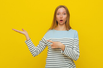 Surprised and amazed girl with fair-haired is laughing and indicating fingers on palm empty space advising novelty product customers, showing advertisement. Indoor studio shot on yellow background