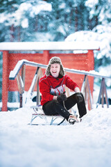 Smiling young cheerful caucasian man is wearing worm red sweatshirt and fun winter ear hat is having fun with sled in a snowy park or forest on winter day. Happy Christmas winter holiday