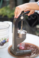 Close up of mechanical iron juicer squeezing pomegranate or tomato juice into glass beaker. Hands of man working on production of natural juice from fresh vegetables and fruits at farmer's market
