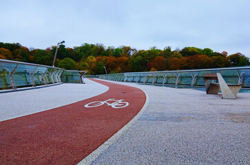 Low-angle view of winding red color bike lane with white bicycle sign on the New Pedestrian Bridge in Kyiv, Ukraine. Beautiful autumn colored trees in the background