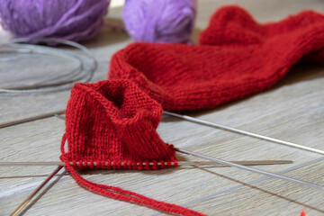 Sock in the process of knitting with needles and yarn of red color on a gray wooden table