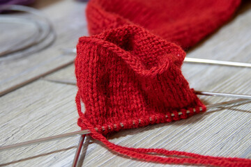 Sock in the process of knitting with needles and yarn of red color on a gray wooden table