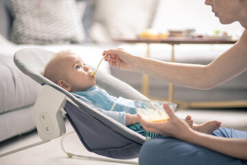Obraz premium Mother spoon feeding her baby boy infant child in baby chair with fruit puree. Baby solid food introduction concept.