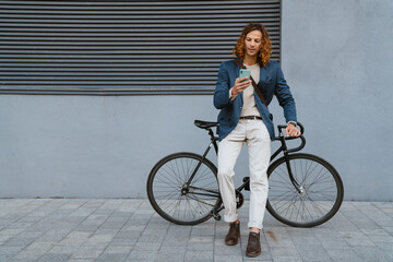 Fototapeta premium Ginger man using mobile phone while walking with bicycle on city street