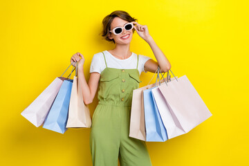 Photo of young cool celebrity woman wear khaki stylish overalls hold new sunglass many bags shopaholic smile isolated on yellow color background - obrazy, fototapety, plakaty