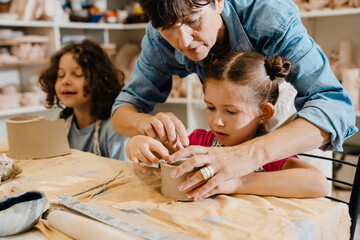 Mature female teacher sculpting with kids and assisting them at ceramics workshop