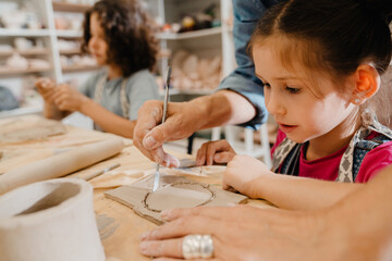 Girl watching teacher hands cutting clay with knife at ceramics workshop