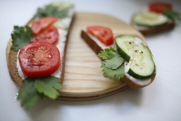 on a wooden kitchen stand are two black bread sandwiches with melted cheese, red tomatoes and green ripe cucumbers and fragrant seasoning and pieces of herbs