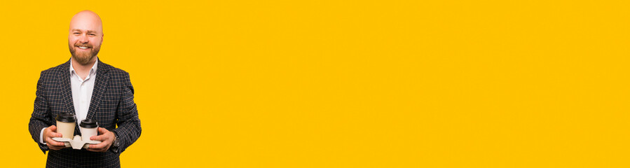 Banner size image of cheerful young bearded man in casual holding two paper cups of coffee over yellow background.