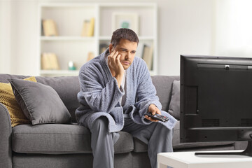 Bored young man in a bathrobe watching tv and sitting on a sofa at home - obrazy, fototapety, plakaty