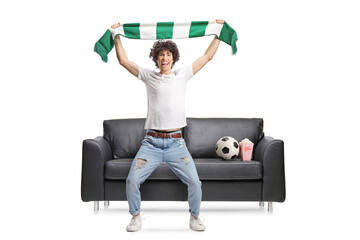 Happy young man cheering with a green and white scarf in front of a couch