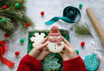 Girl's hands in red knitted sweater put gingerbread in the form of snowman into gift box with Christmas tree and snowflake gingerbreads next to pine branch, ribbons and wrapping paper. Christmas vibes