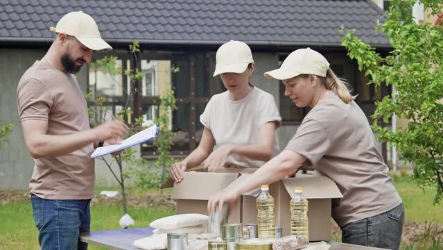 Young members of charity organization pack donations boxes with canned and non-perishable products outdoors. Charity, donation and volunteering concept, global food crisis