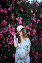 Pregnant girl on the background of pink roses with a hat on her head. Red lipstick on the lips