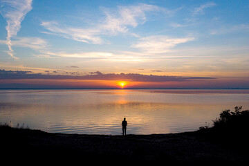 drone view of a dark silhouette of a woman standing on the edge of the sea with a colourful sunset in front of her; autumn sunset over the baltic sea, hel peninsula, poland - obrazy, fototapety, plakaty