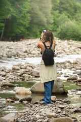 a young tourist woman with beautiful wavy hair, with a backpack on her shoulders, travels in the mountains near the river and takes pictures of nature on the phone