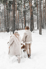 Love romantic young couple girl, guy in snowy cold winter forest walking with pet, dog of hunting breed russian borzoi. Sighthound, wolfhound owner. Having fun, laughing. Stylish fur coat, woolen hat