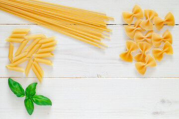 Maccheroni raw, spaghetti, farfalle, pipe, rigatoni, penne, rigate, leaf of basil, on white wooden board background, top view, space to copy text..