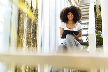 An african american woman with big curly hair sitting in a staircase in garden house smiling and writing in a folder.