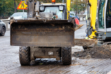 A wide metal bucket of a road bulldozer is raised above the surface of the road section being repaired.