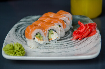 Appetizing portion of sushi rolls philadelphia with red fish salmon, with soft cheese, cucumbers, avocado and ginger with wasabi, on a gray plate on a black table, on background of yellow fruit juice