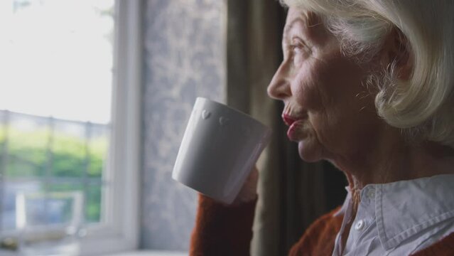 Senior woman sitting by radiator at home trying to keep warm during energy and cost of living crisis drinking hot drink and shivering - shot in slow motion 