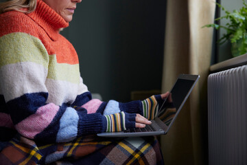 Woman In Gloves With Laptop Trying To Keep Warm By Radiator During Cost Of Living Energy Crisis