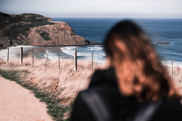 blonde caucasian girl with backpack and black coat out of focus from back walking on dirt road leading to quiet lonely beach, sandfly bay, new zealand - Powered by Adobe