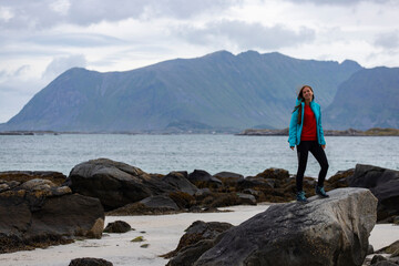 Fototapeta premium girl in blue jacket walks on the beach with huge rocks and mountains in the background, the Lofoten islands in norway and their harsh landscapes